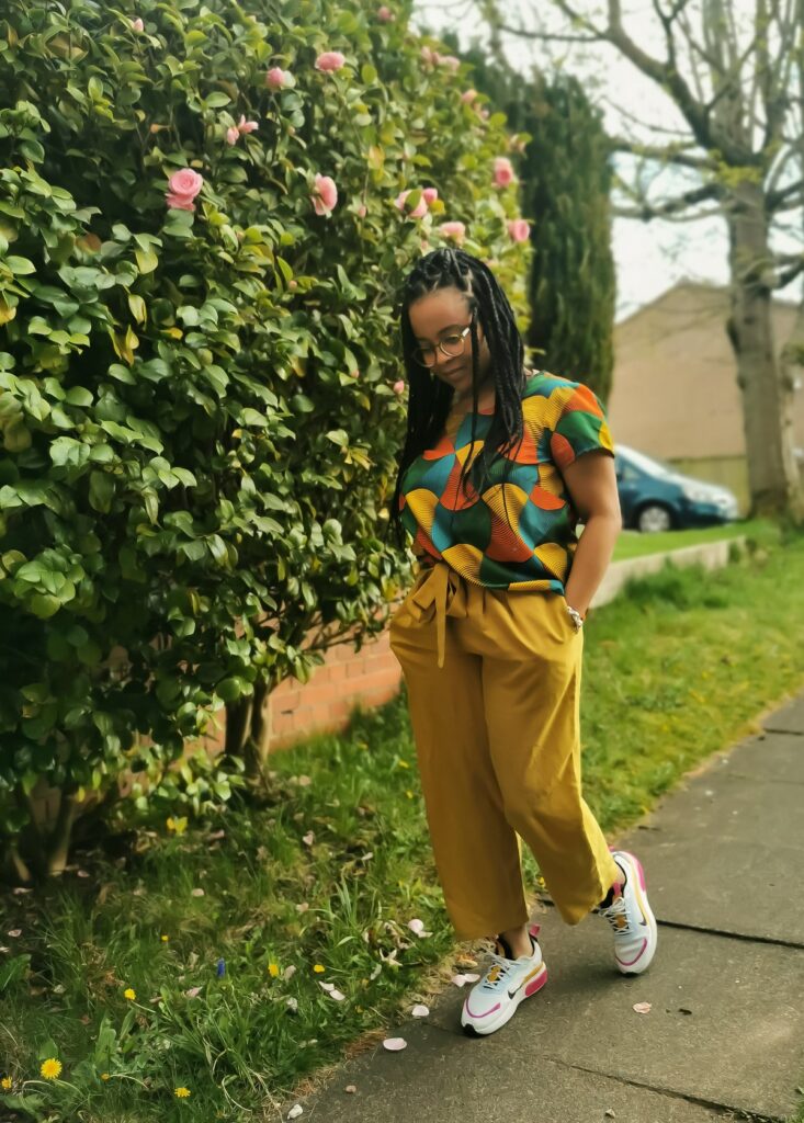 Black woman with braids stands outdoors wearing a colourful Scout Tee and handmade trousers. She is looking down towards the ground and is in mid stride